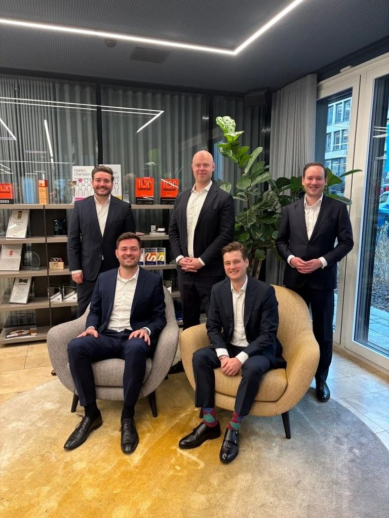 Group photo of five men in business suits, two seated and three standing in a modern office with shelves and plants, representing consulting leadership team collaboration