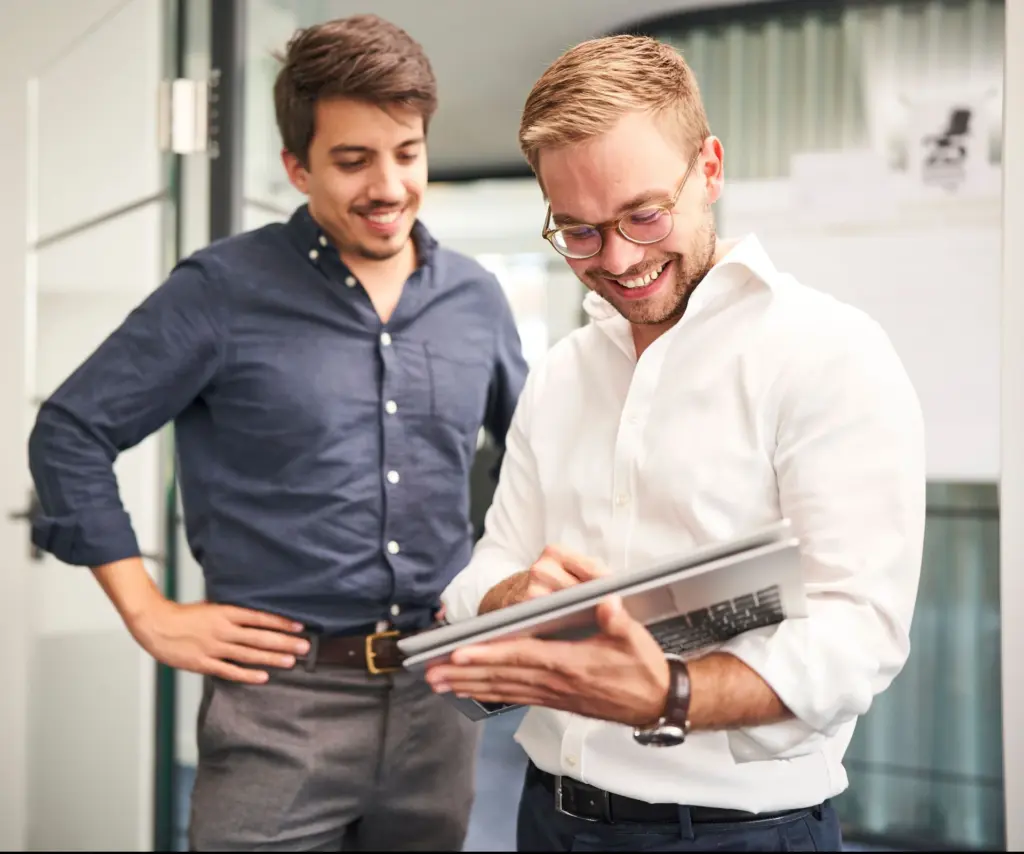 Two men in an office, one smiling and holding a tablet while the other stands behind with hands on hips, both wearing smart casual attire, illustrating collaborative discussion and consulting