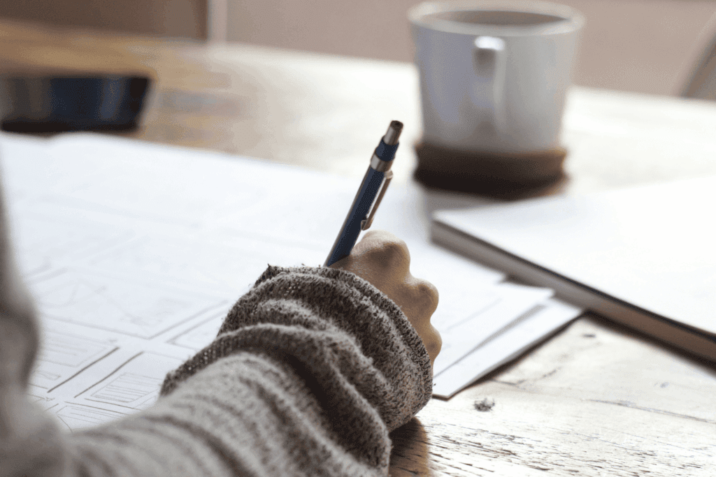 Person writing notes on paper with a pen next to a coffee mug on a wooden desk | H&Z
