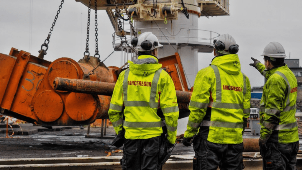 Three MacGregor workers in high-visibility jackets observing crane equipment at a shipyard | H&Z