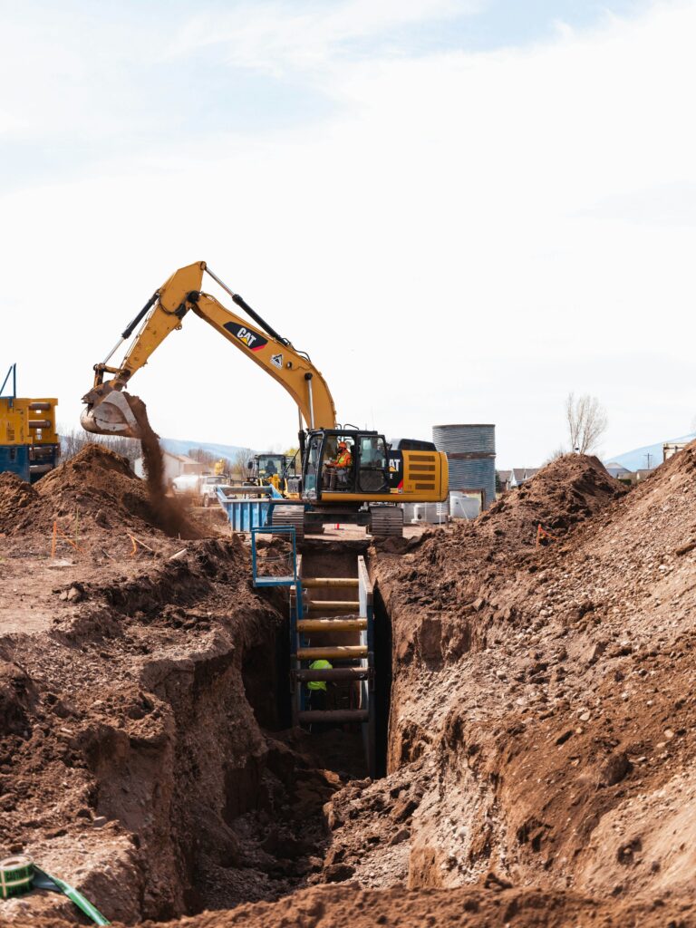 Yellow CAT excavator digging a deep shored trench on a construction site under a bright sky | H&Z